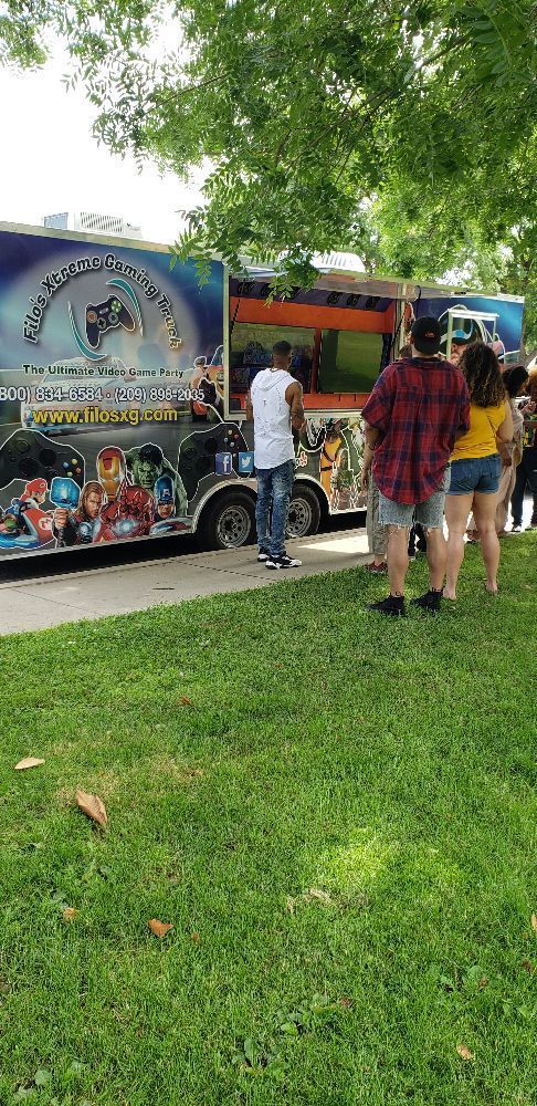 People stand in line by a colorful trailer in a grassy area, under trees.