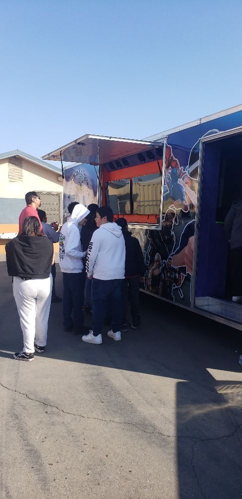 People gather at a colorful food truck under a blue sky, outside a building with a white wall.