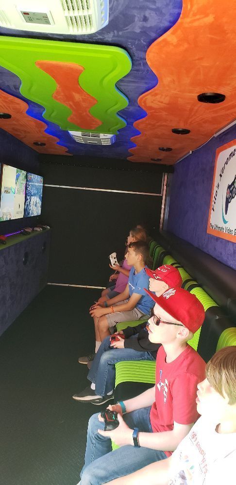 Children playing video games inside a colorful van; they hold controllers and watch screens.