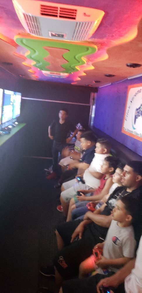 Inside a gaming truck, several people sit, playing games. Screens line one side, with a man standing nearby.