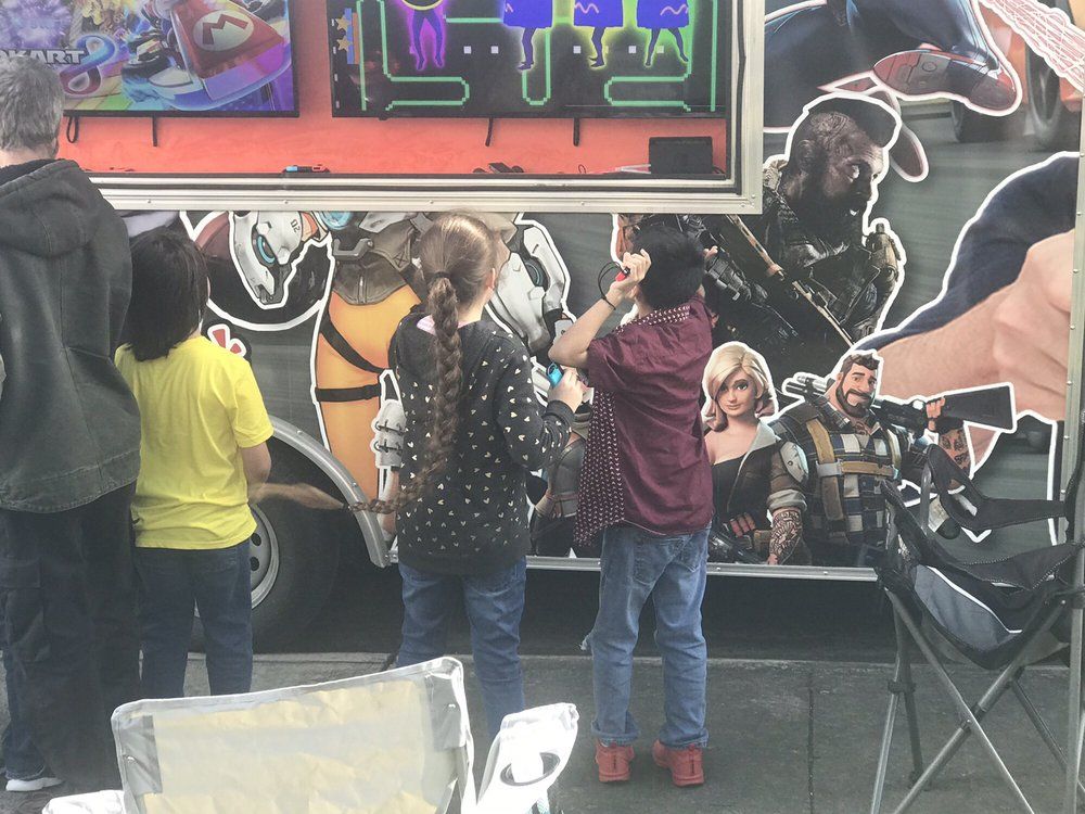 People playing a video game at an outdoor event, standing in front of a game-themed truck.