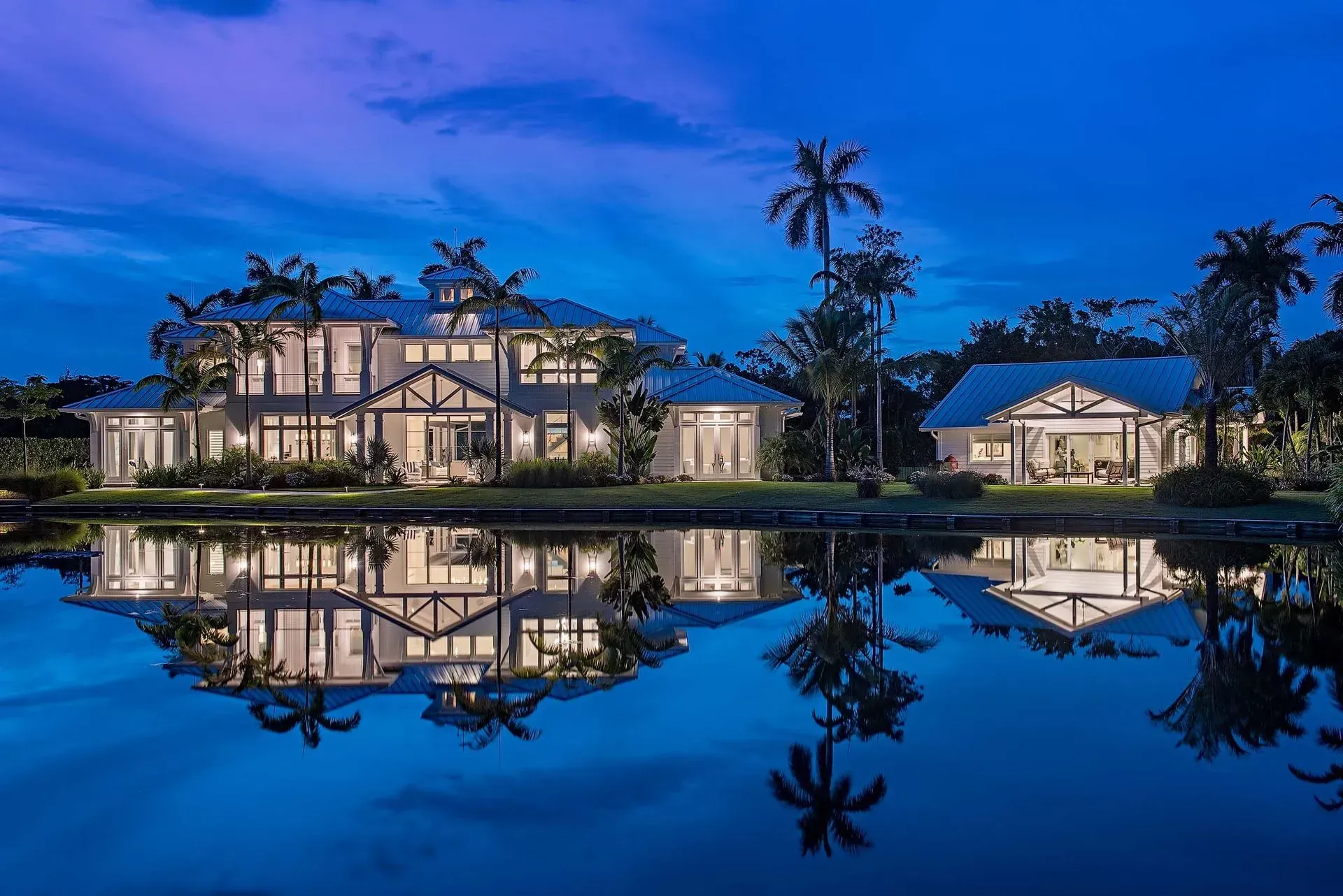 Large waterfront home with reflections in water at dusk, palm trees, illuminated by interior lights.