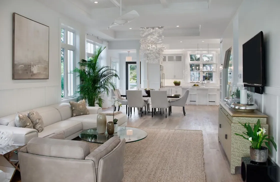 Bright, airy living room with white walls, sectional sofa, dining table, and kitchen in the background.