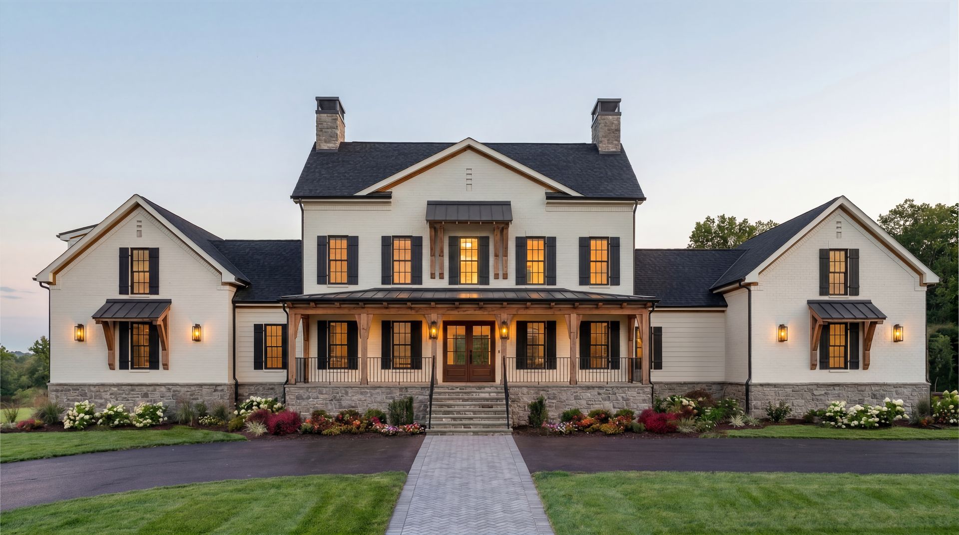Large modern farmhouse with gray roof, white siding, warm-lit windows, and a front lawn at dusk
