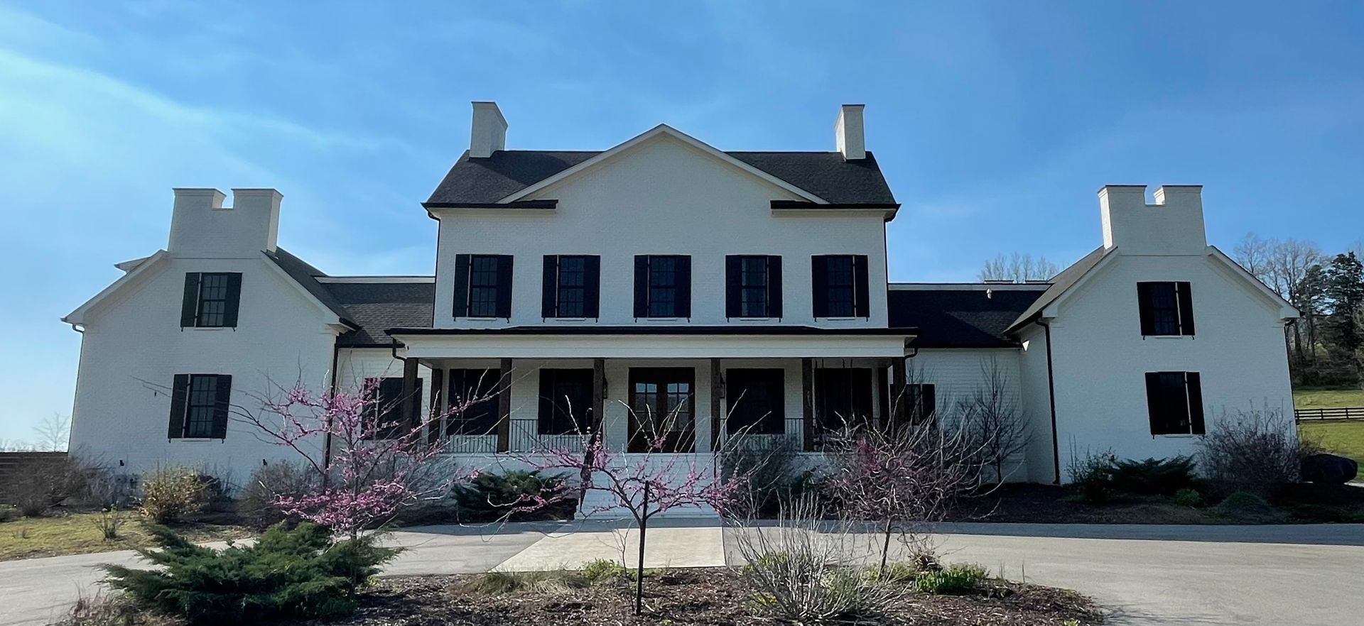 Large white two-story house with dark shutters and a front porch, seen behind flowering shrubs