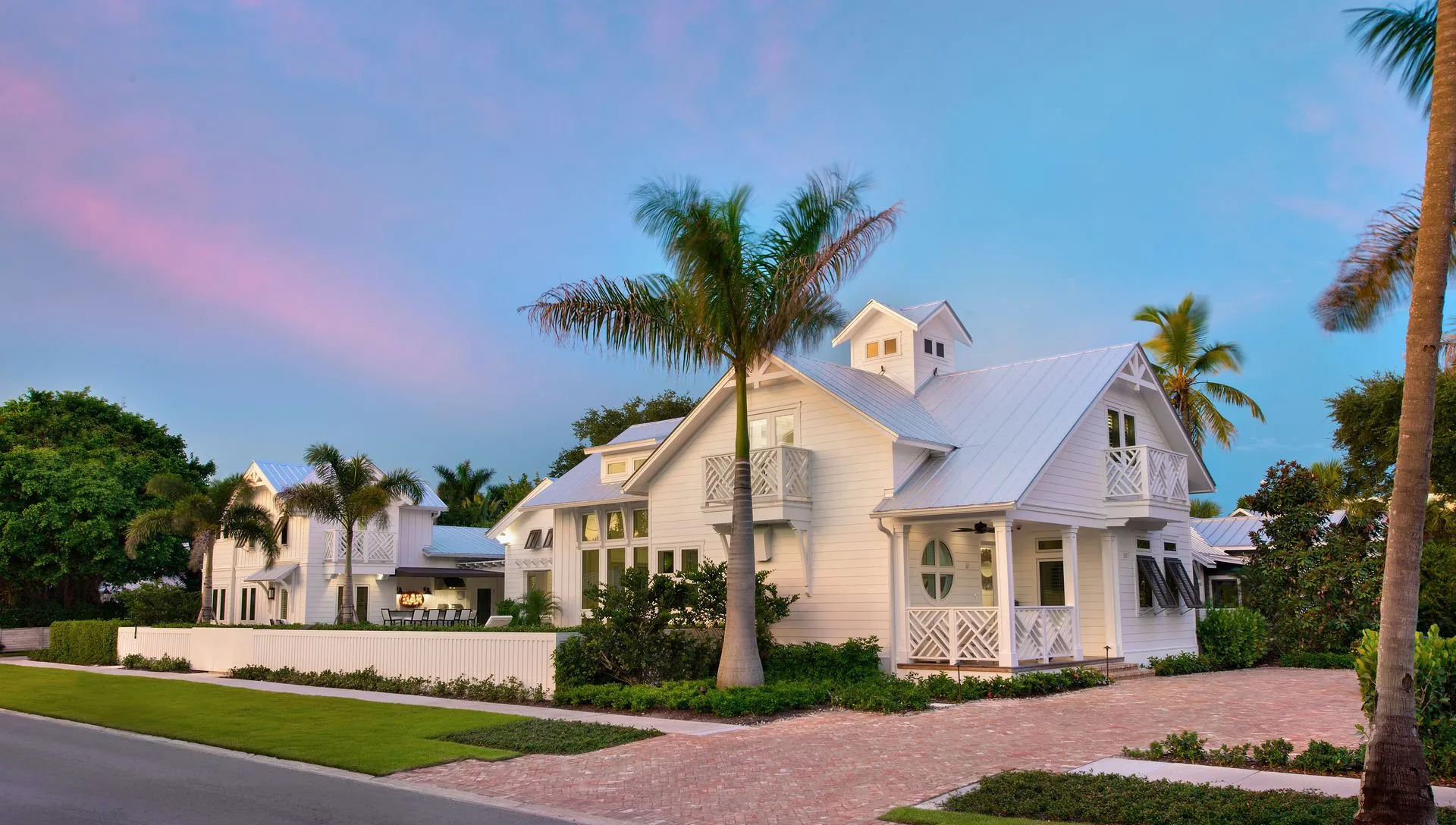 White house with a metal roof, palm trees, and brick driveway under a dusk sky.