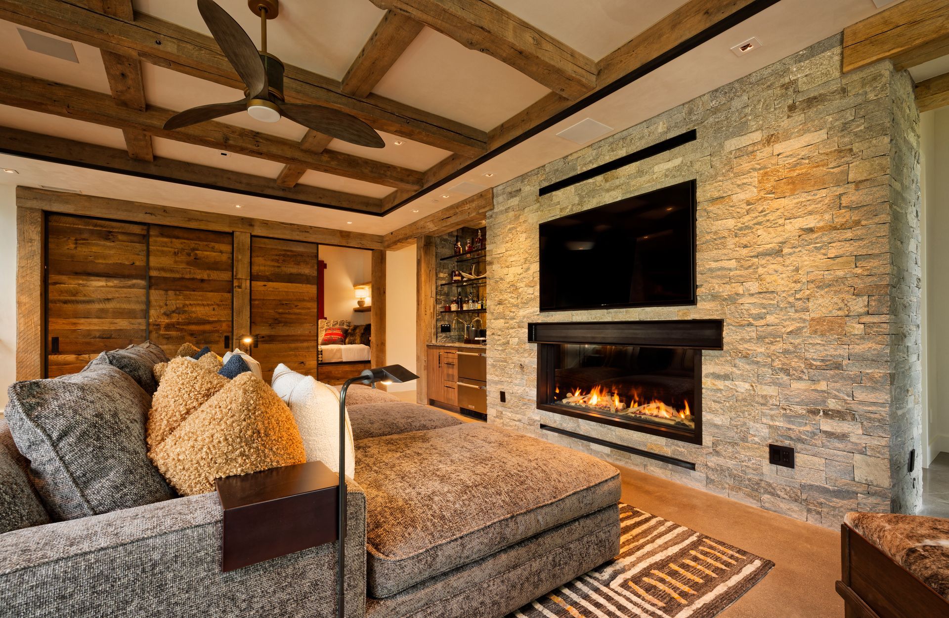 Cozy living room with stone fireplace, wall-mounted TV, sectional sofa, and exposed wood beams