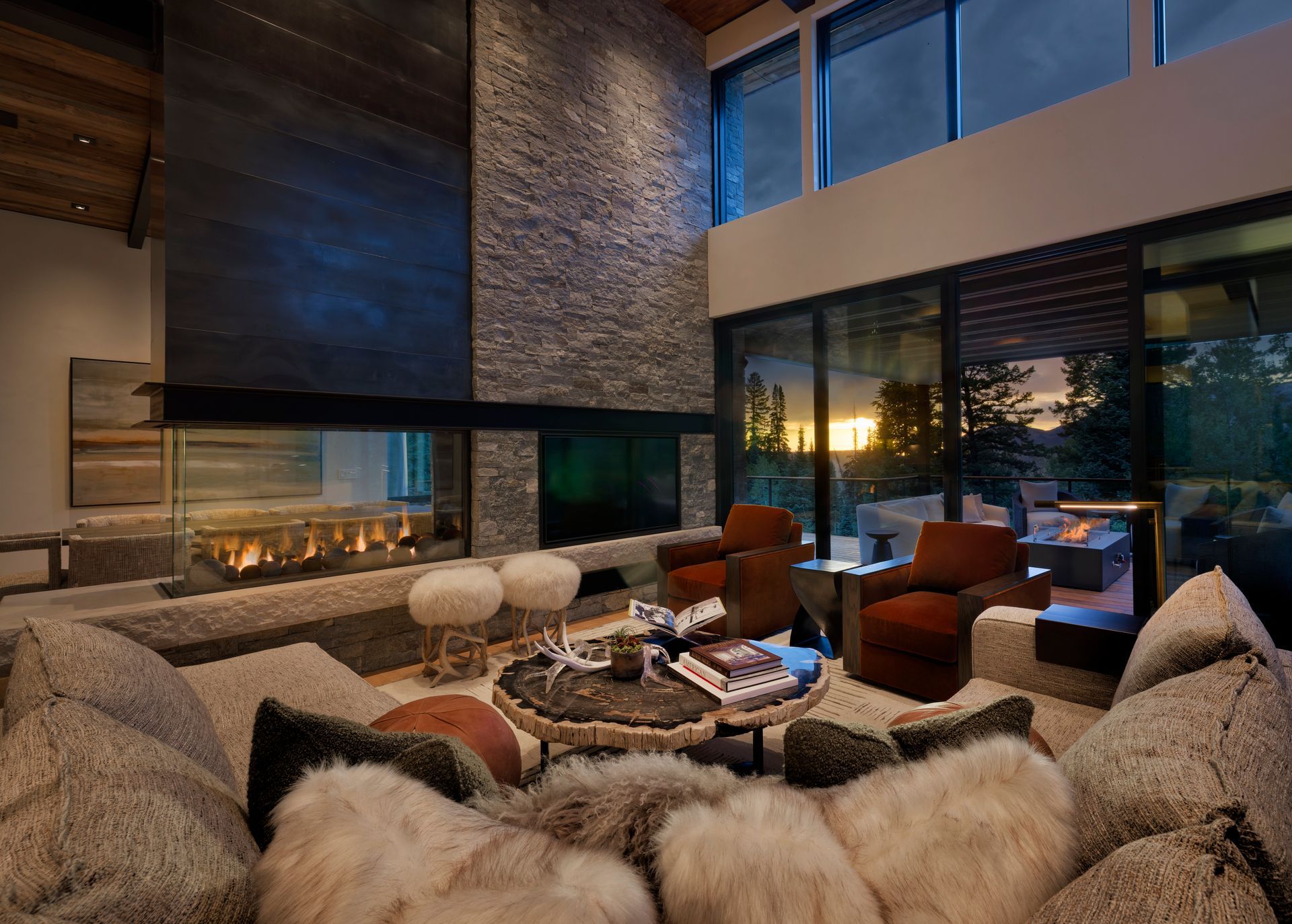 Modern living room with stone fireplace, leather chairs, fur sofa, and large windows at sunset