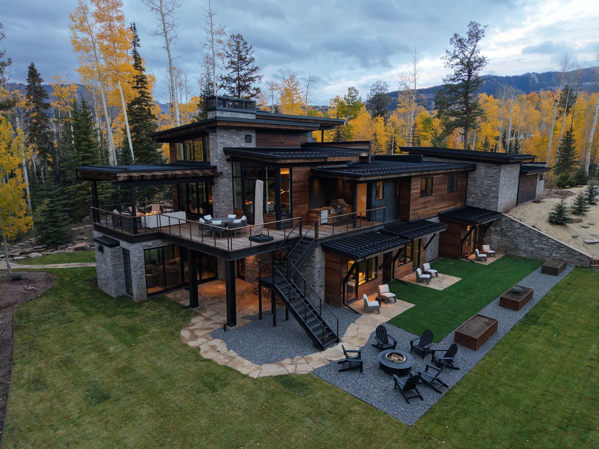 Modern rustic mountain lodge with dark wood decks and stone walls, surrounded by autumn trees and outdoor seating.