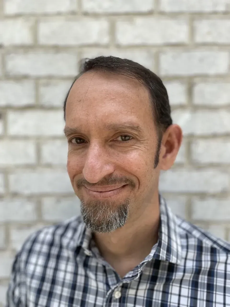 Man with goatee smiles, wearing a checkered shirt, in front of a white brick wall.