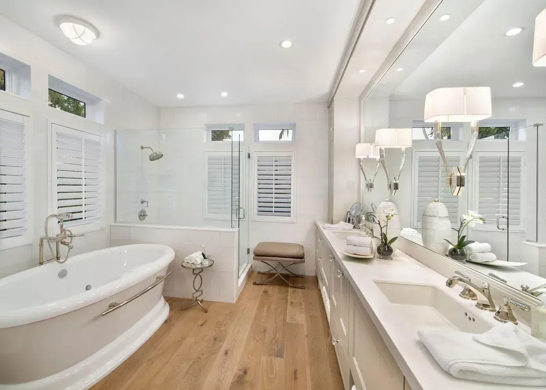Elegant white bathroom with a tub, shower, and vanity. Wooden floors, bright lighting, and white shutters.