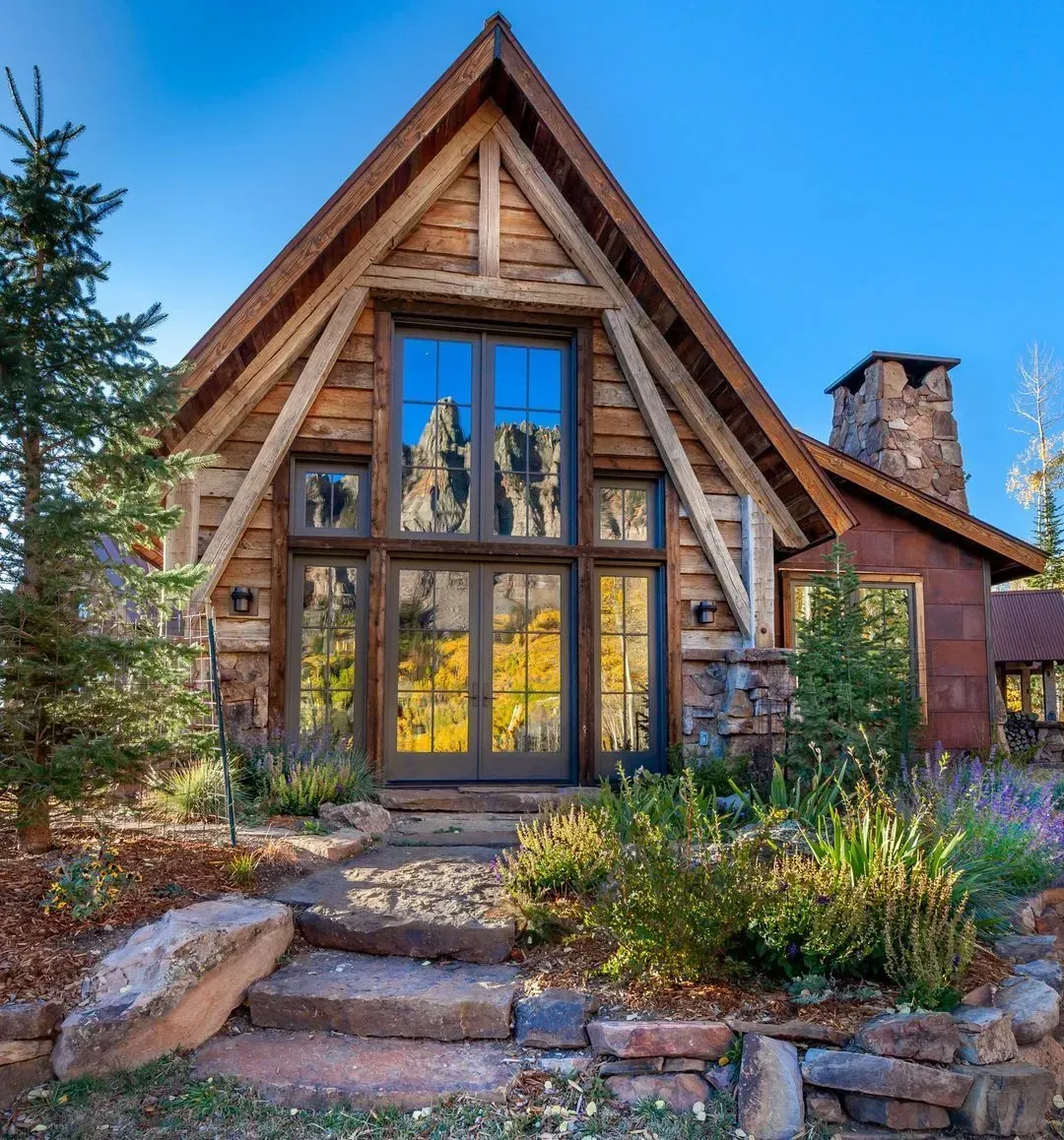 Rustic cabin with wooden exterior, stone chimney, and large windows reflecting fall foliage.