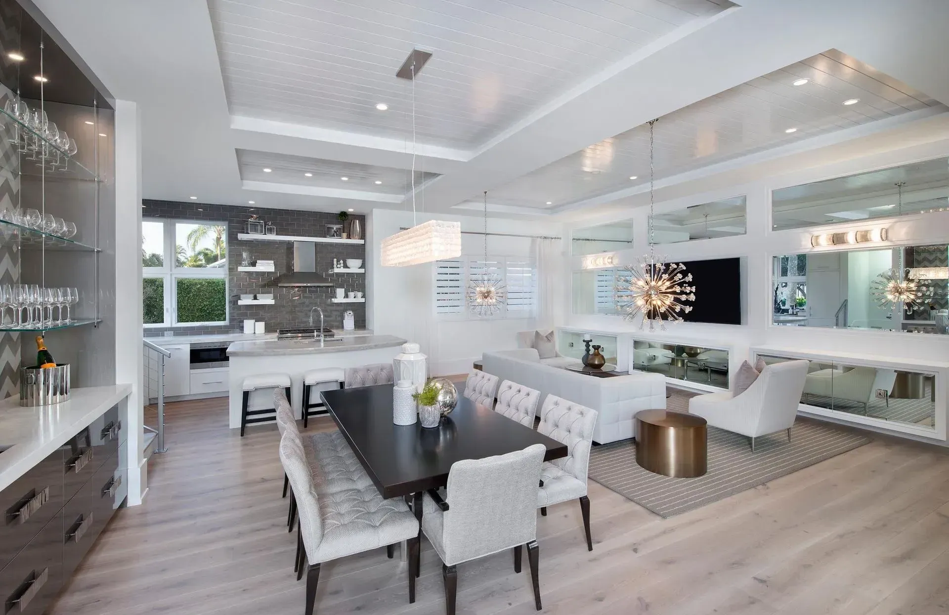Modern open-concept living space with dining area, kitchen, and seating area; light wood floors, mirrors, and white furniture.