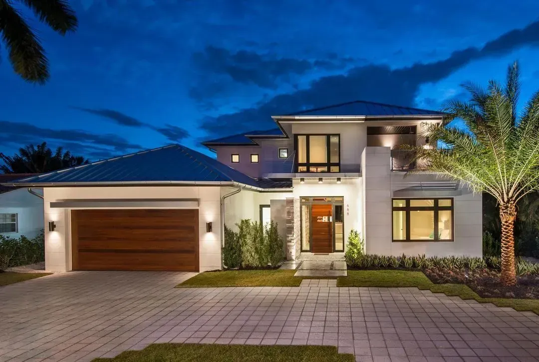 Modern two-story house with a white exterior, wooden garage door, and palm tree lit at night under a blue sky.