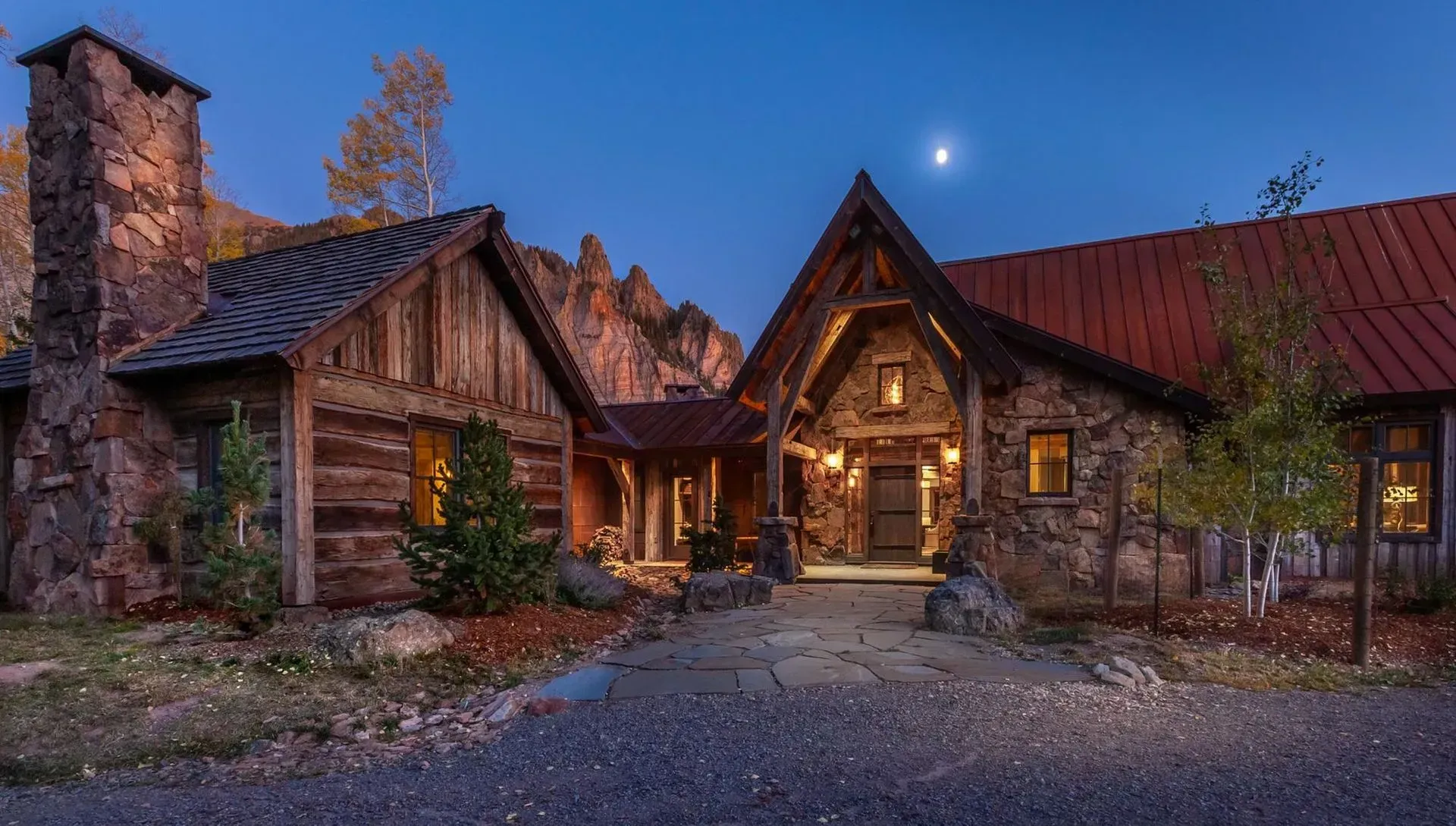 Rustic stone and wood lodge at dusk, lit interior, mountain backdrop, moon in the sky.