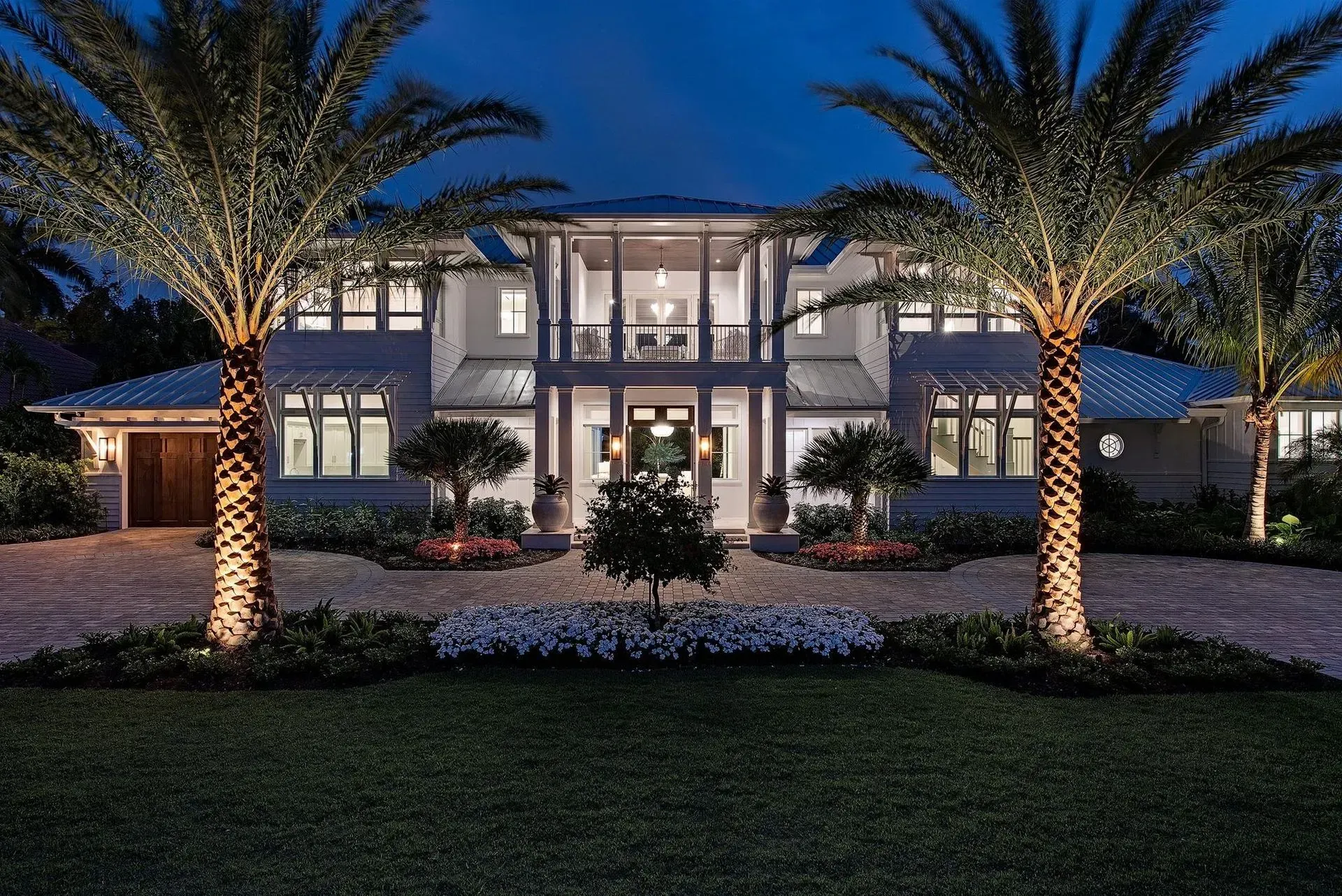 Luxury home at night with palm trees illuminated. Driveway and landscaping visible.