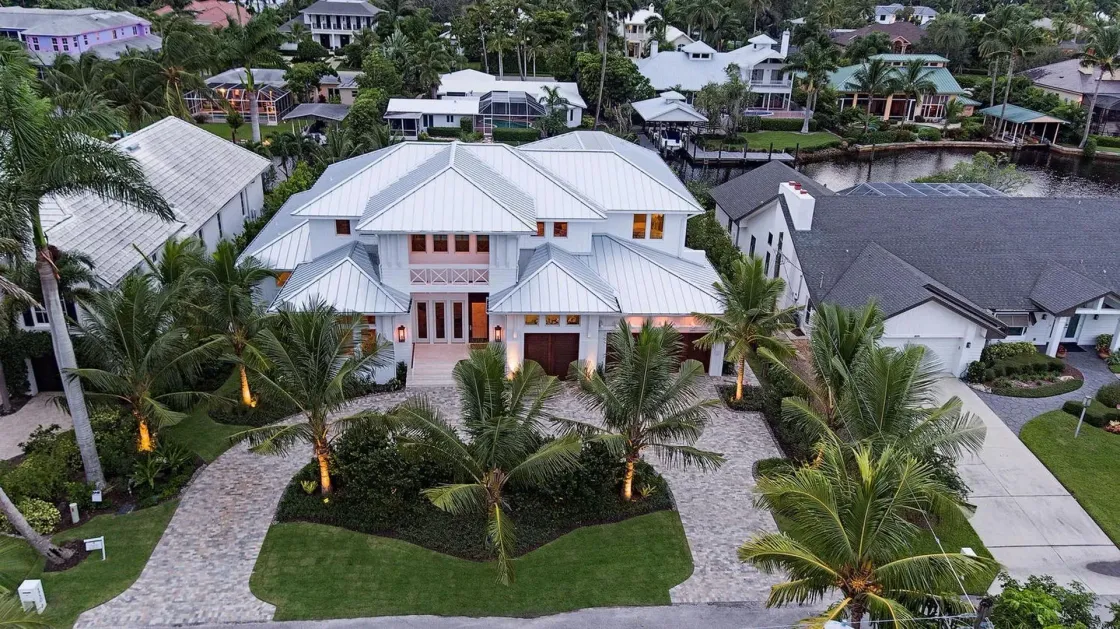 Aerial view of a white multi-story house with a white roof, palm trees, and a driveway in a residential area.