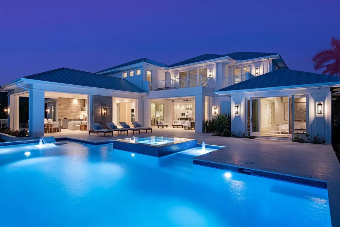 Luxury home with pool at night, illuminated blue; lounge chairs, open-air living spaces, water fountains.
