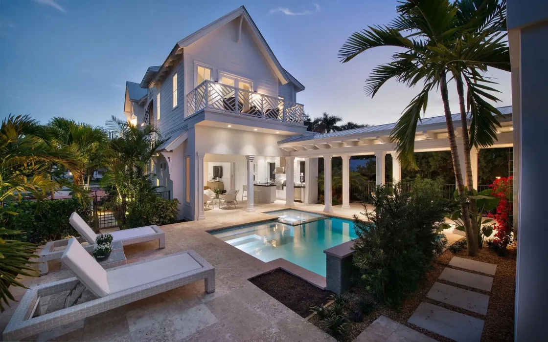 White house with a pool and palm trees at dusk; lounge chairs on the patio.