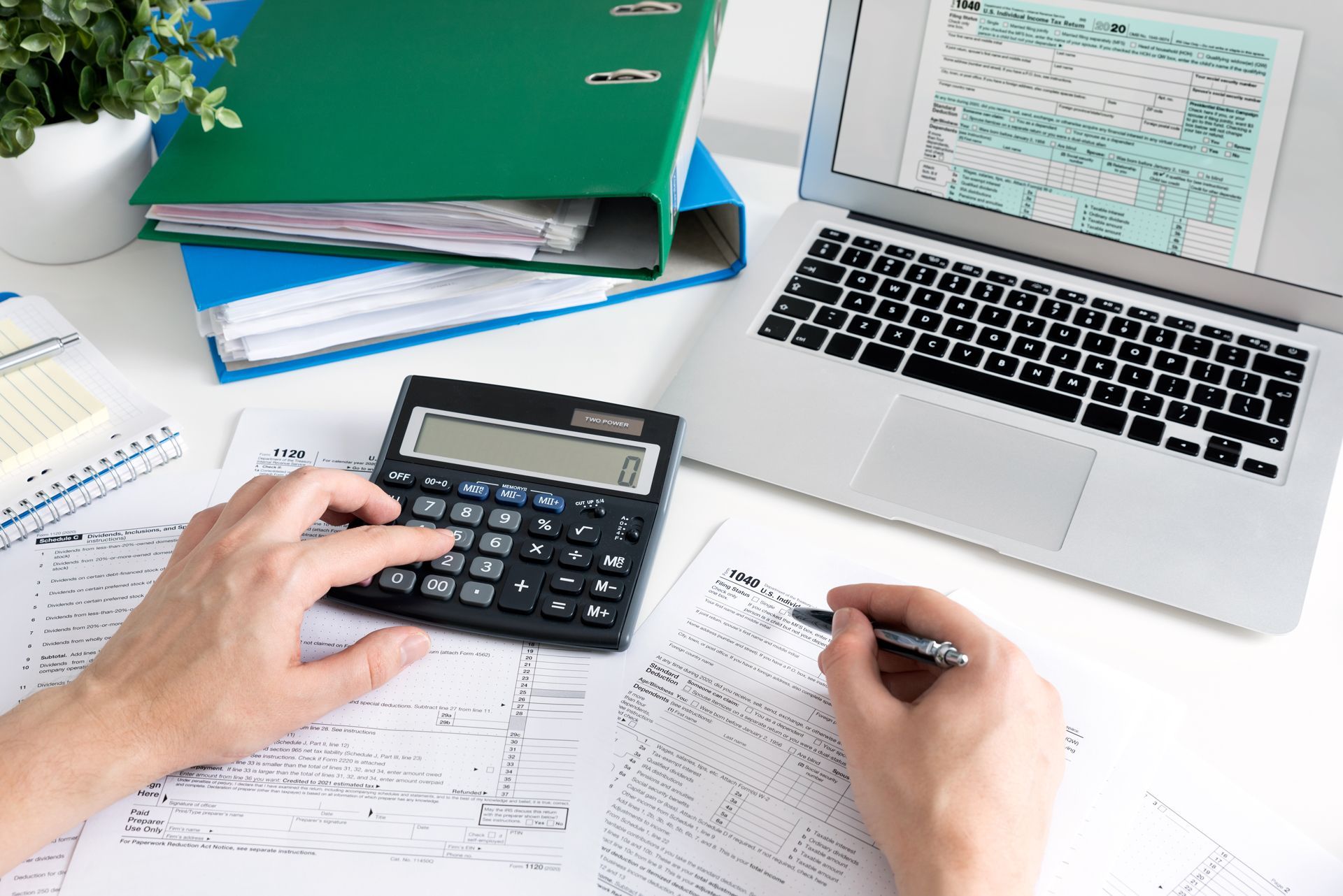 Hands using a calculator over paperwork beside a laptop and office folders on a desk
