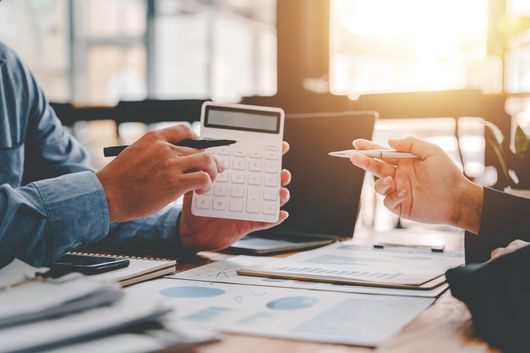 Two people discussing finances at a desk, using a calculator and pointing at paperwork in a bright office.