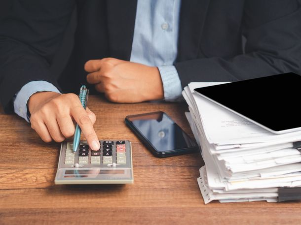 Person using a calculator at a desk with a phone and a stack of papers and a tablet nearby