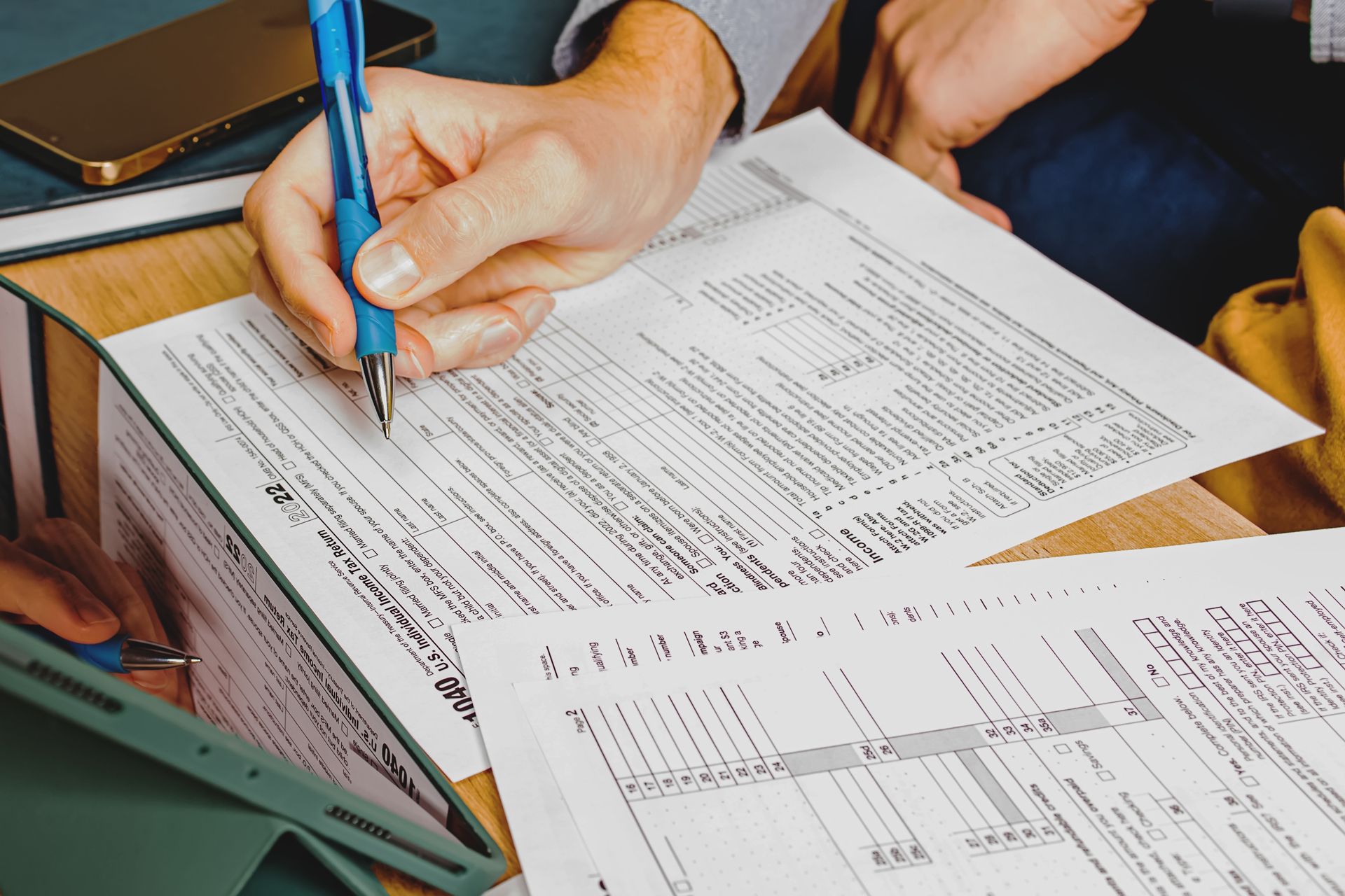 Hands writing on printed forms with a blue pen at a desk, surrounded by paperwork and a tablet