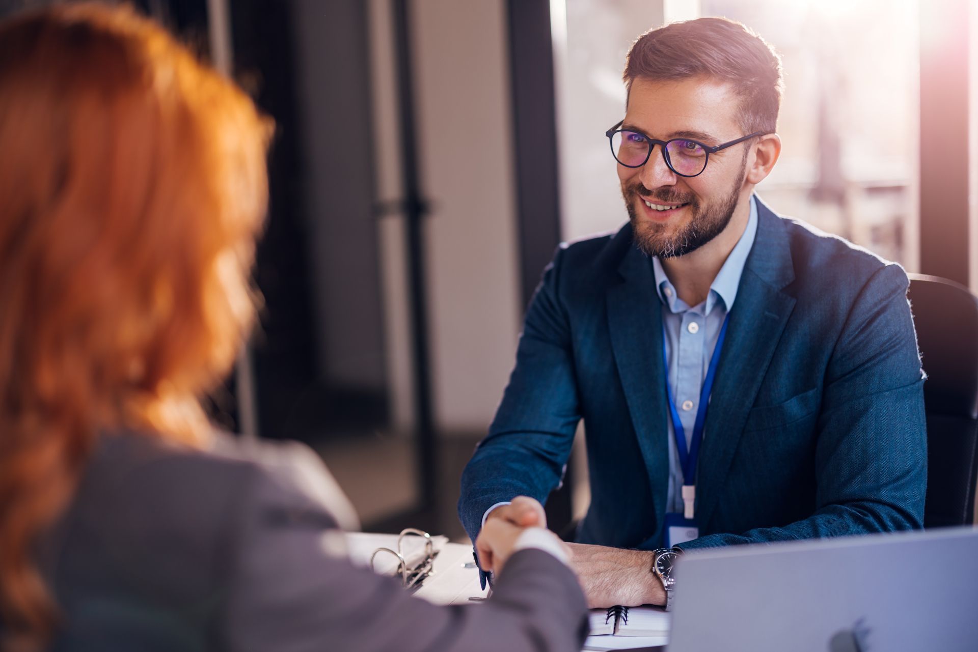 Two people shaking hands across a desk in a bright office, one smiling in a blue blazer