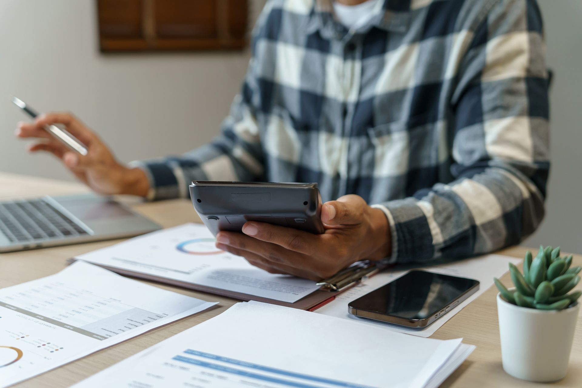 Person in plaid shirt using a calculator at a desk with laptop, papers, phone, and a small plant.