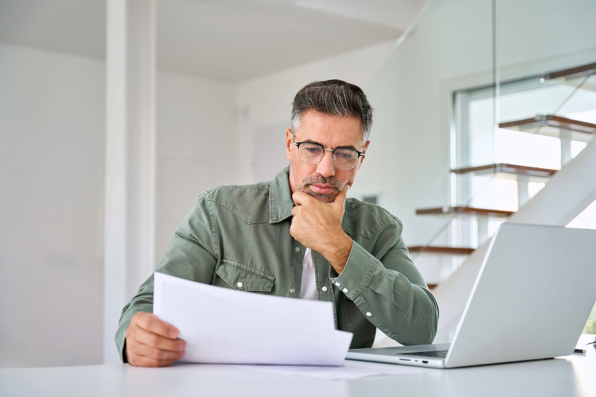 Man reviewing paperwork at a desk with a laptop in a bright office