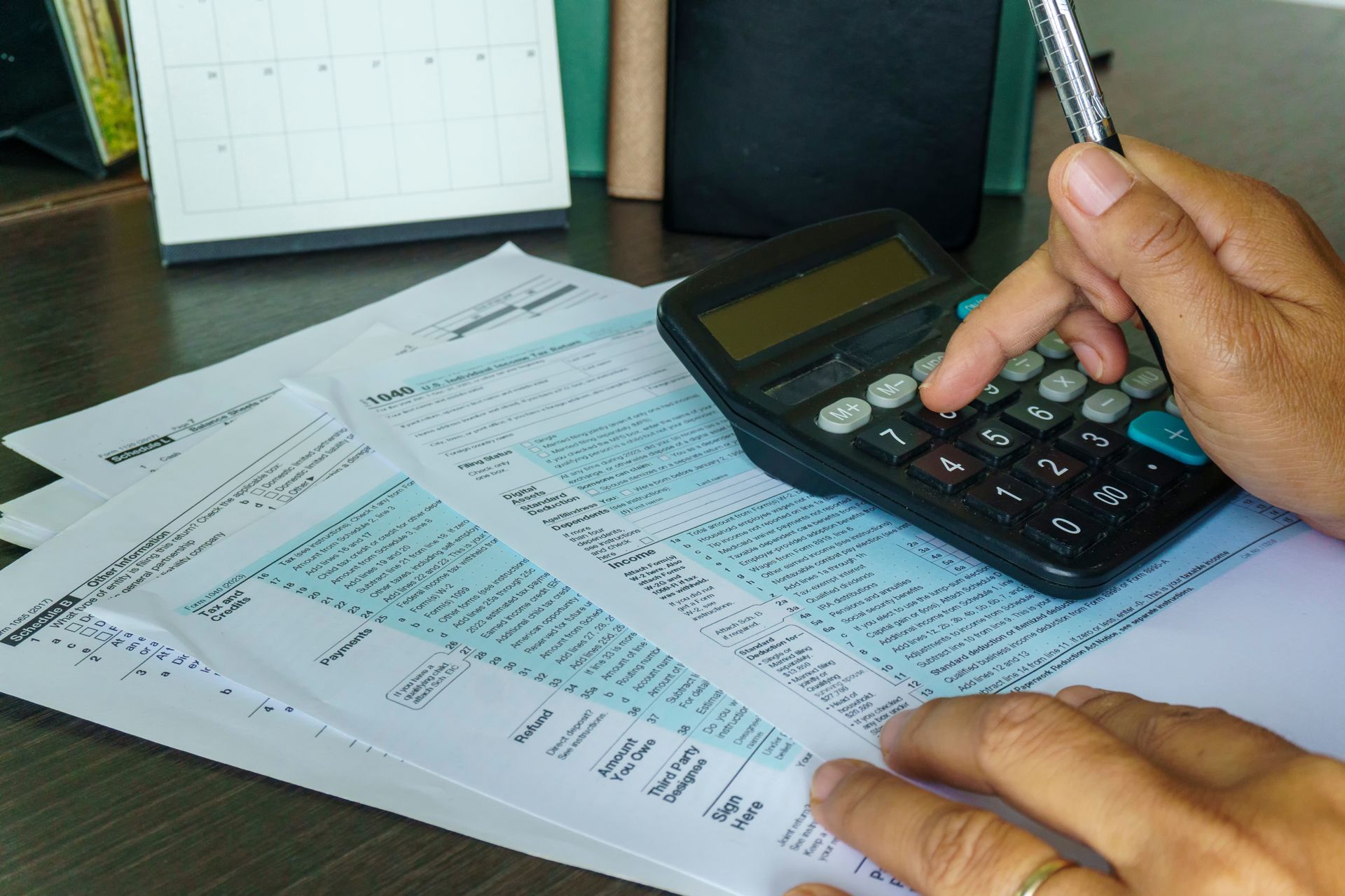 Hands using a calculator on paper bills and forms at a desk with a calendar and pen holder