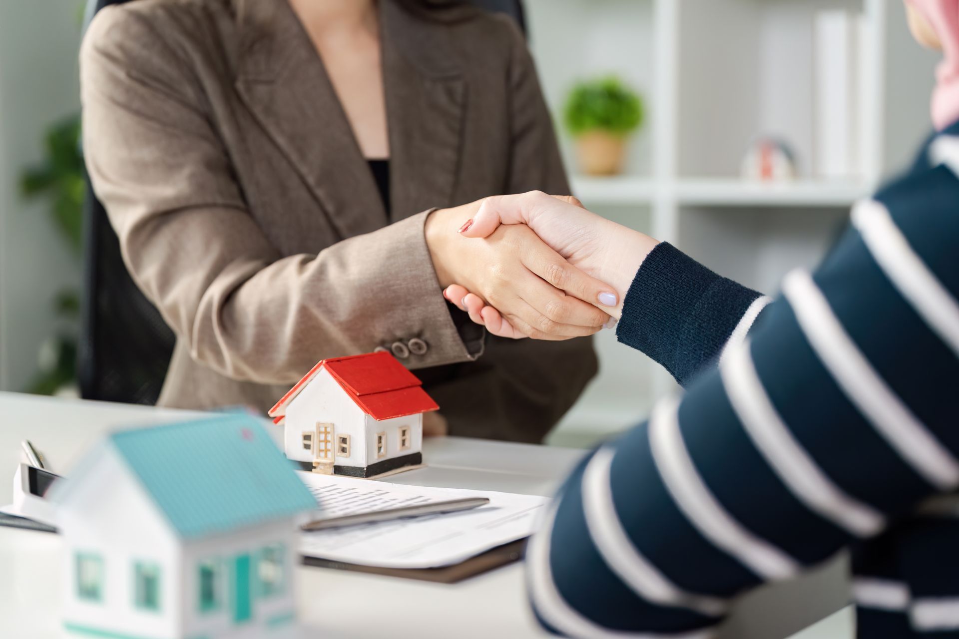 Two people shake hands across a desk with small house models and paperwork, suggesting a real estate deal