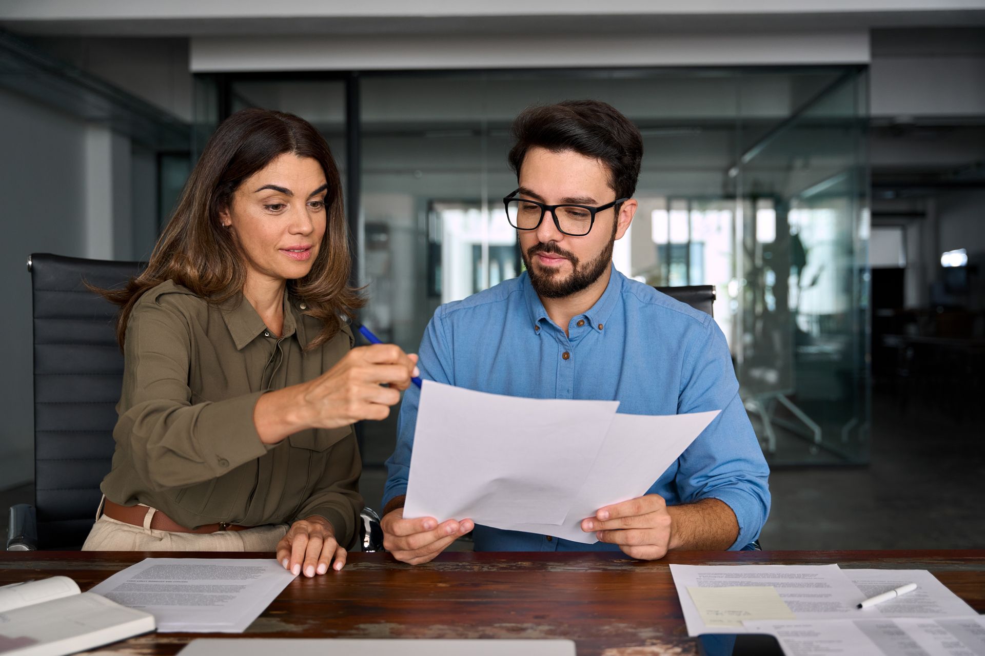 Two coworkers reviewing a document at a desk in a modern office, one pointing at the page.