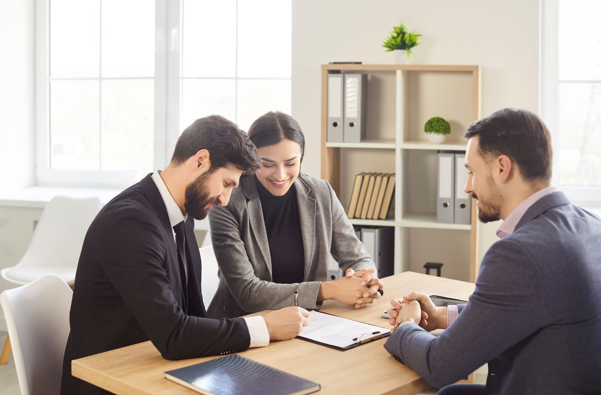 Three coworkers in business attire reviewing documents at a table in a bright office