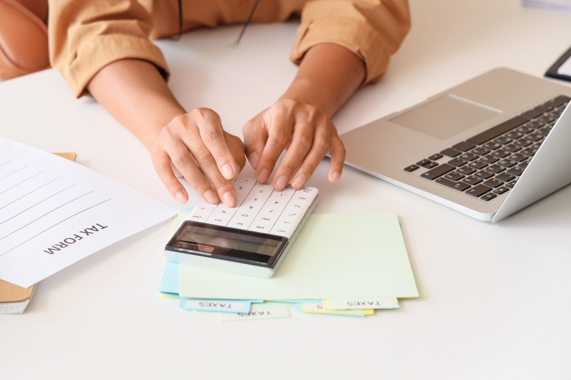 Hands typing on a calculator beside a laptop and documents on a white desk