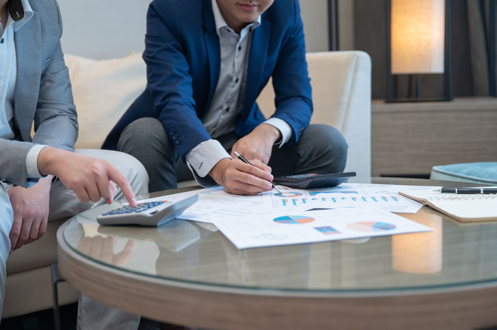 Two people in business attire reviewing charts and a calculator on a glass coffee table in an office meeting