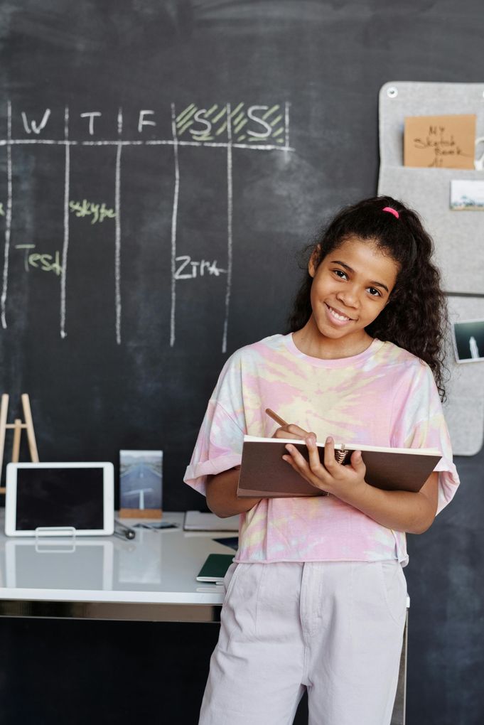 A young girl is standing in front of a blackboard holding a notebook and a pen.