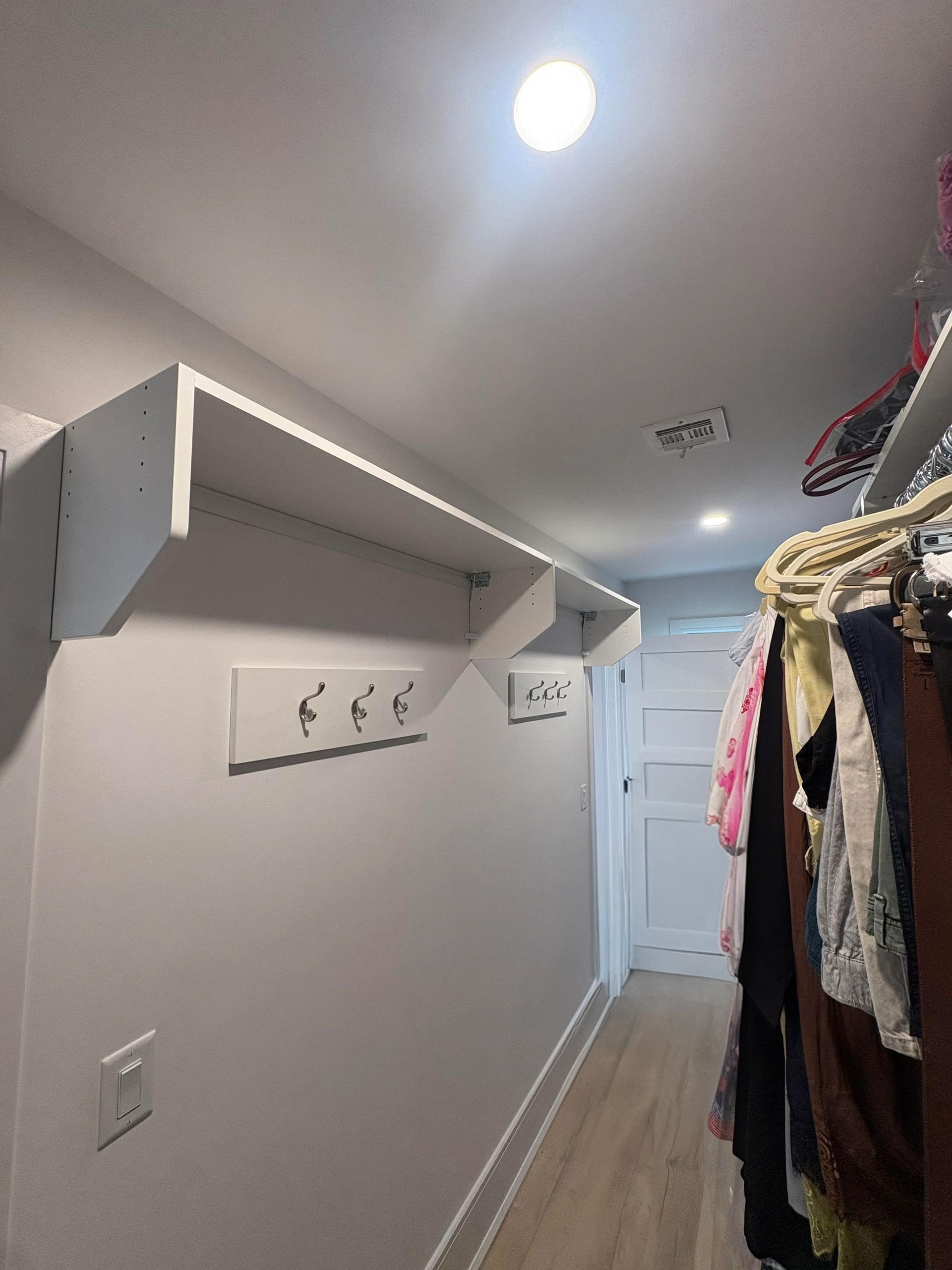Narrow hallway closet with hanging clothes, wall shelves, and a white door at the end.