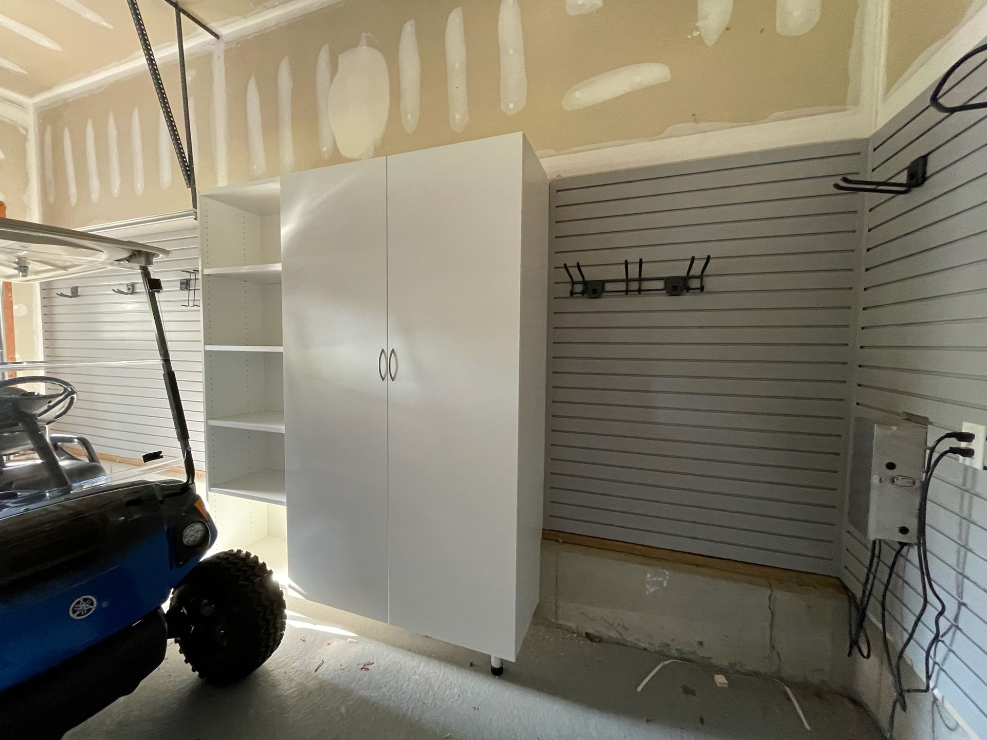 A garage interior featuring a tall white cabinet, adjacent open shelving, and wall-mounted storage tracks on gray walls.