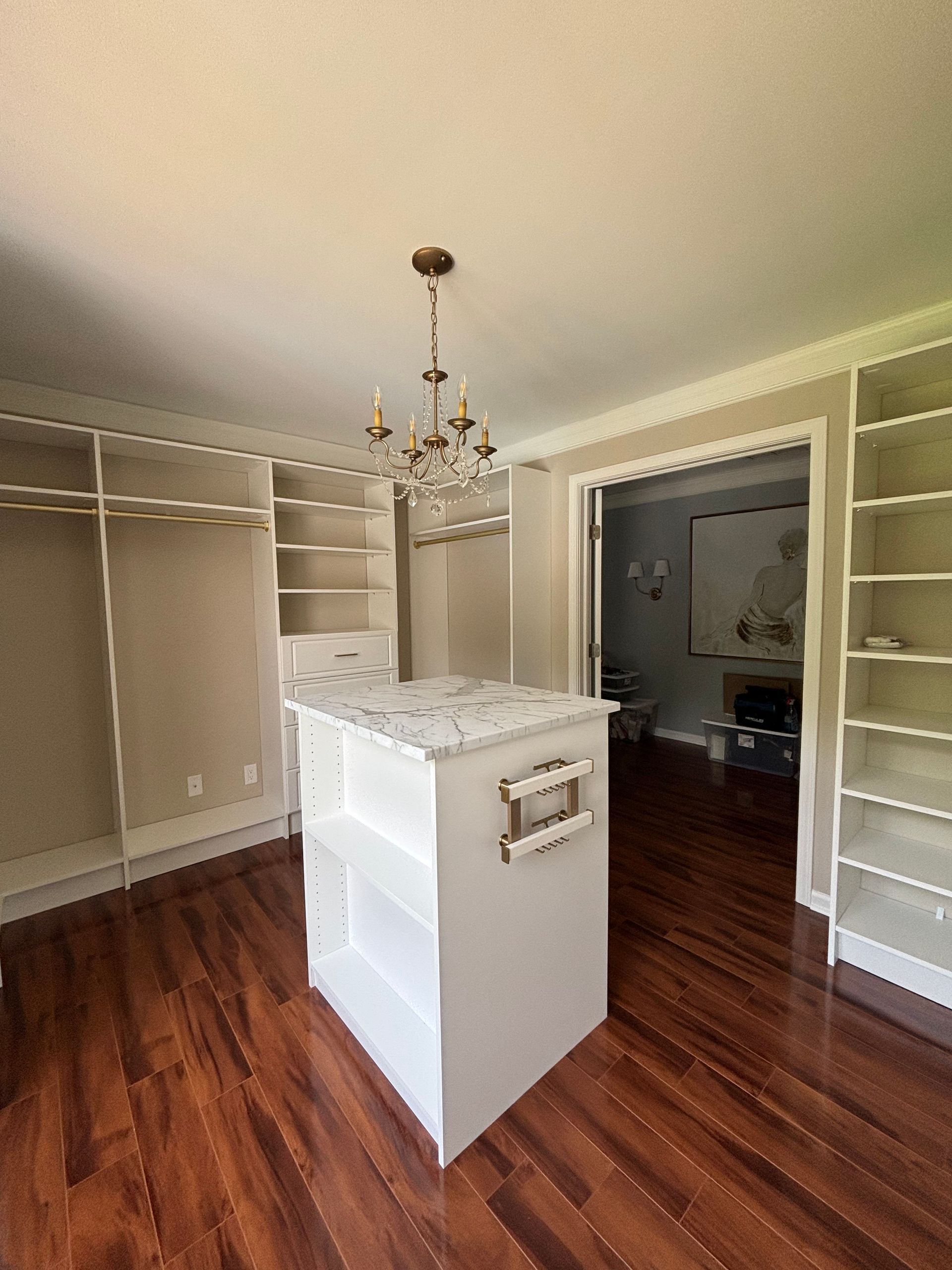 Empty walk-in closet with hardwood floors, white built-in shelving, and a small island under a chandelier.