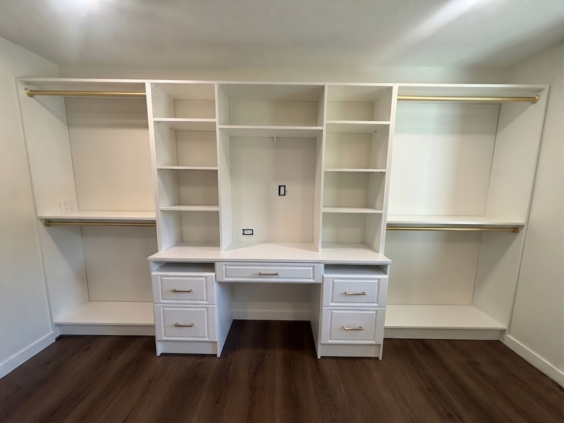 White built-in desk with shelves and drawers in a room, framed by wall cabinets and wood flooring