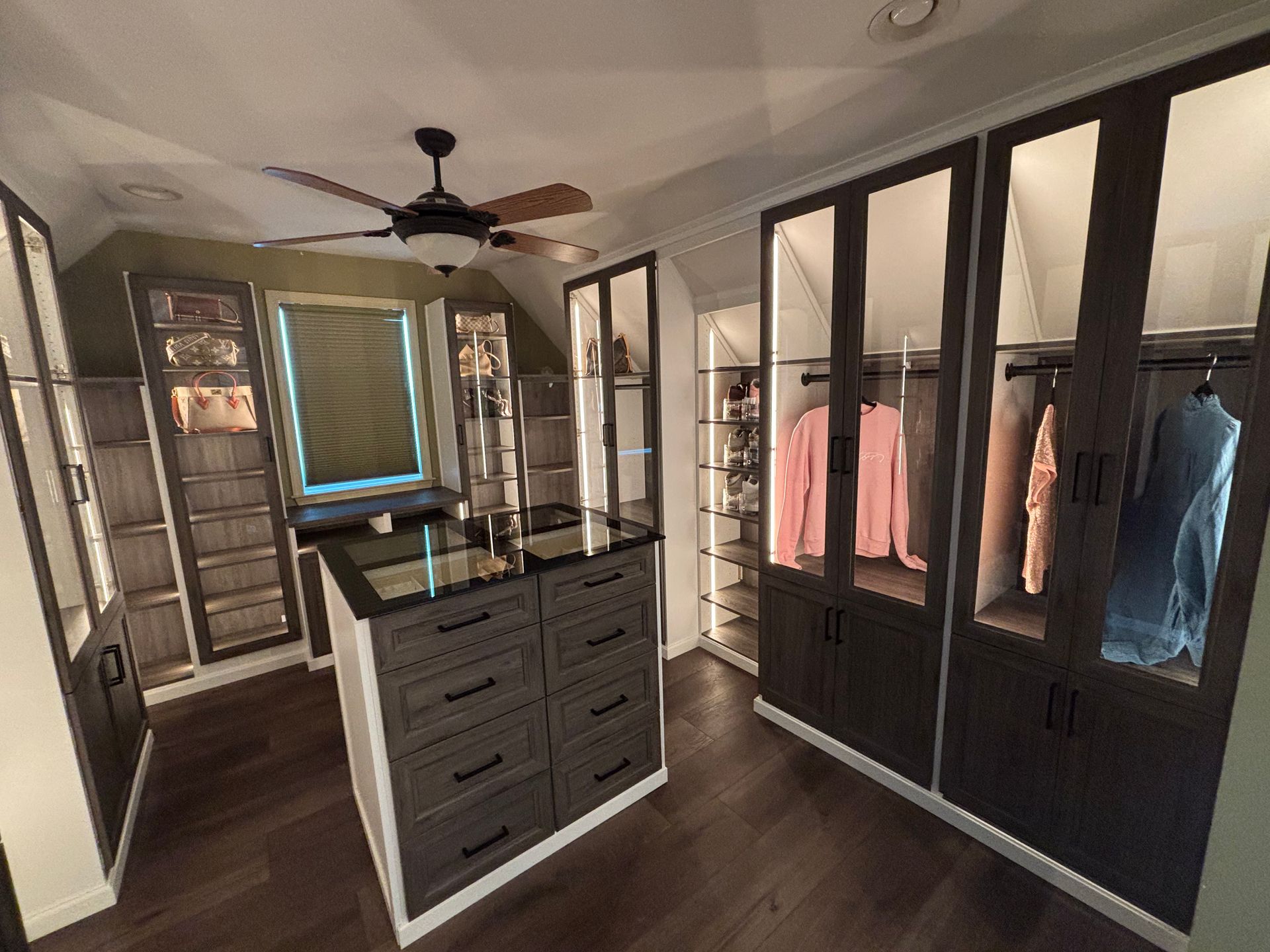 A modern, walk-in closet featuring dark cabinetry with glass doors, a center island with drawers, and a ceiling fan.