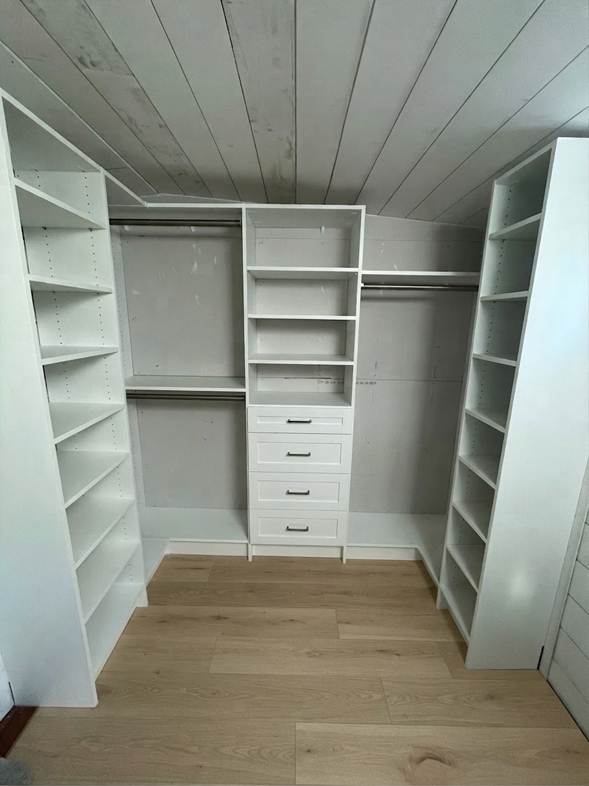 Empty walk-in closet with white shelves, drawers, and hanging rods, plus wood flooring and slanted ceiling.
