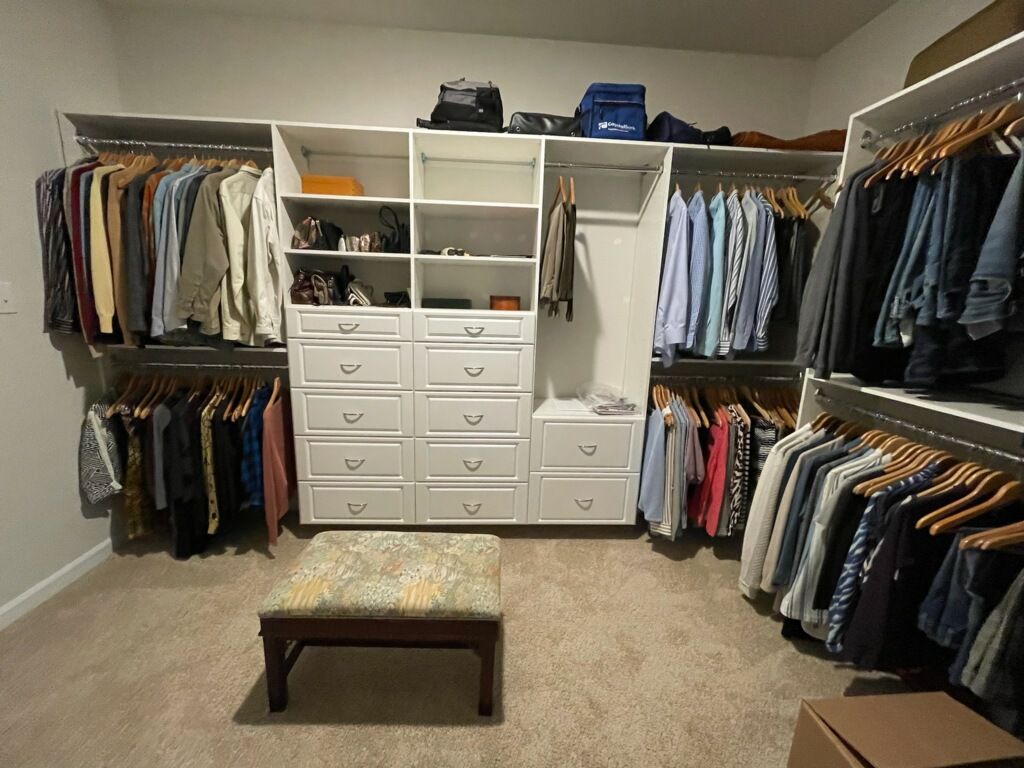 A walk-in closet with white shelving, hanging clothes, drawers, and a central upholstered stool on a carpeted floor.