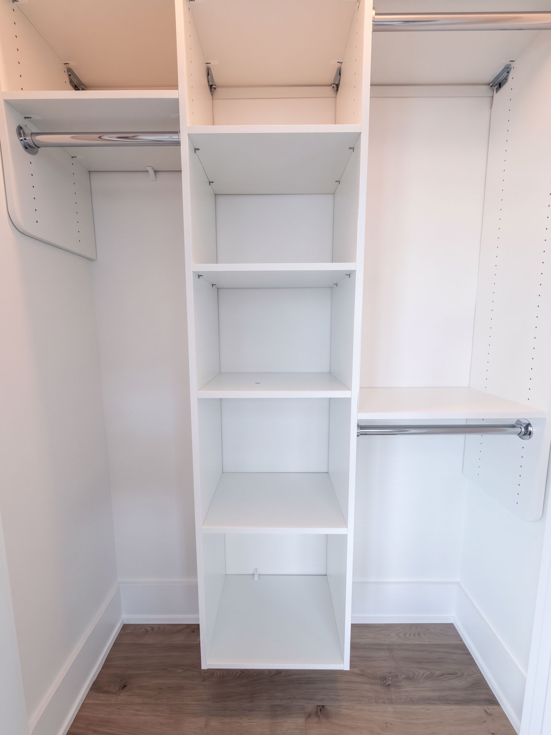 Empty walk-in closet with white built-in shelves and hanging rods on both sides, wood floor, and central shelving unit