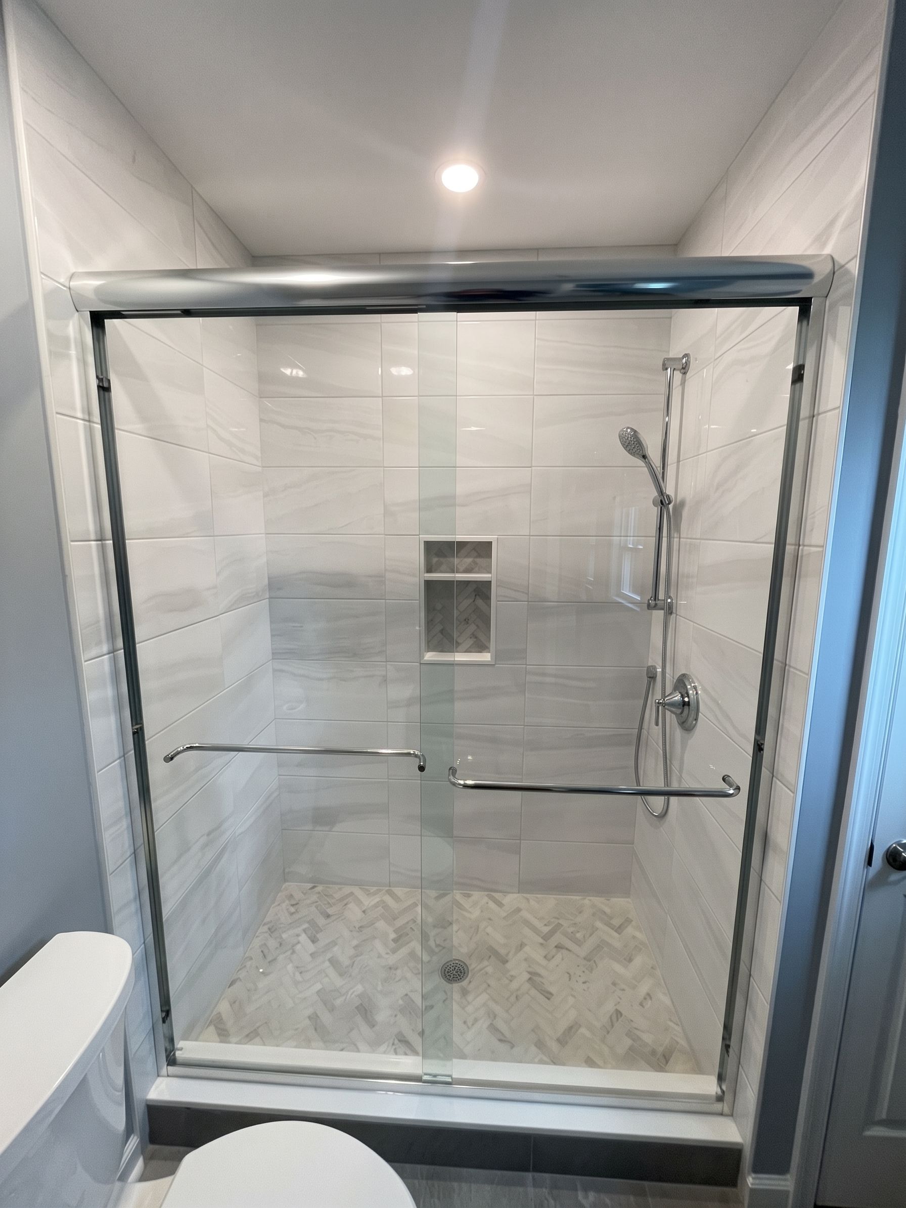 Modern glass shower stall with tiled walls, built-in niche, grab bar, and marble-patterned floor.