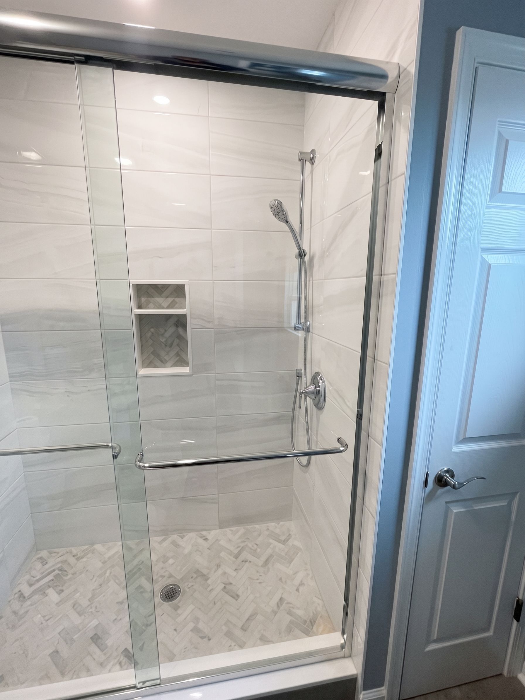 Glass shower with white tile, built-in niche, and chrome grab bar next to a bathroom door.