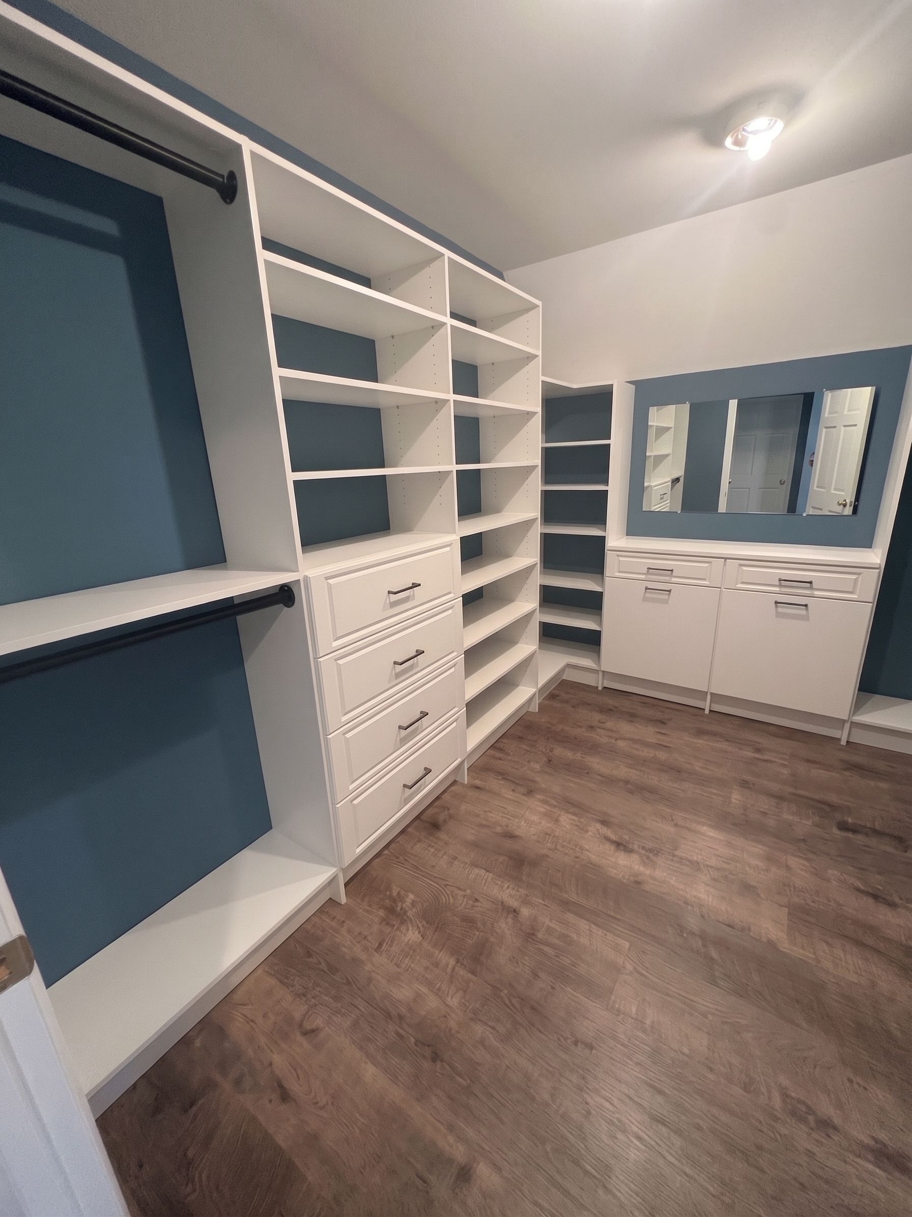 walk-in closet with white built-in shelves and drawers, blue walls, wood flooring, and a vanity cabinet.