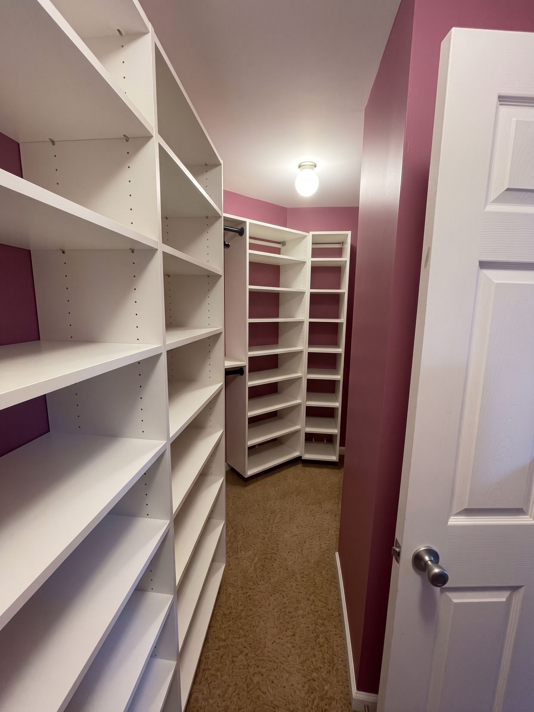 Narrow walk-in closet with white shelving units, maroon walls, and carpeted floor.