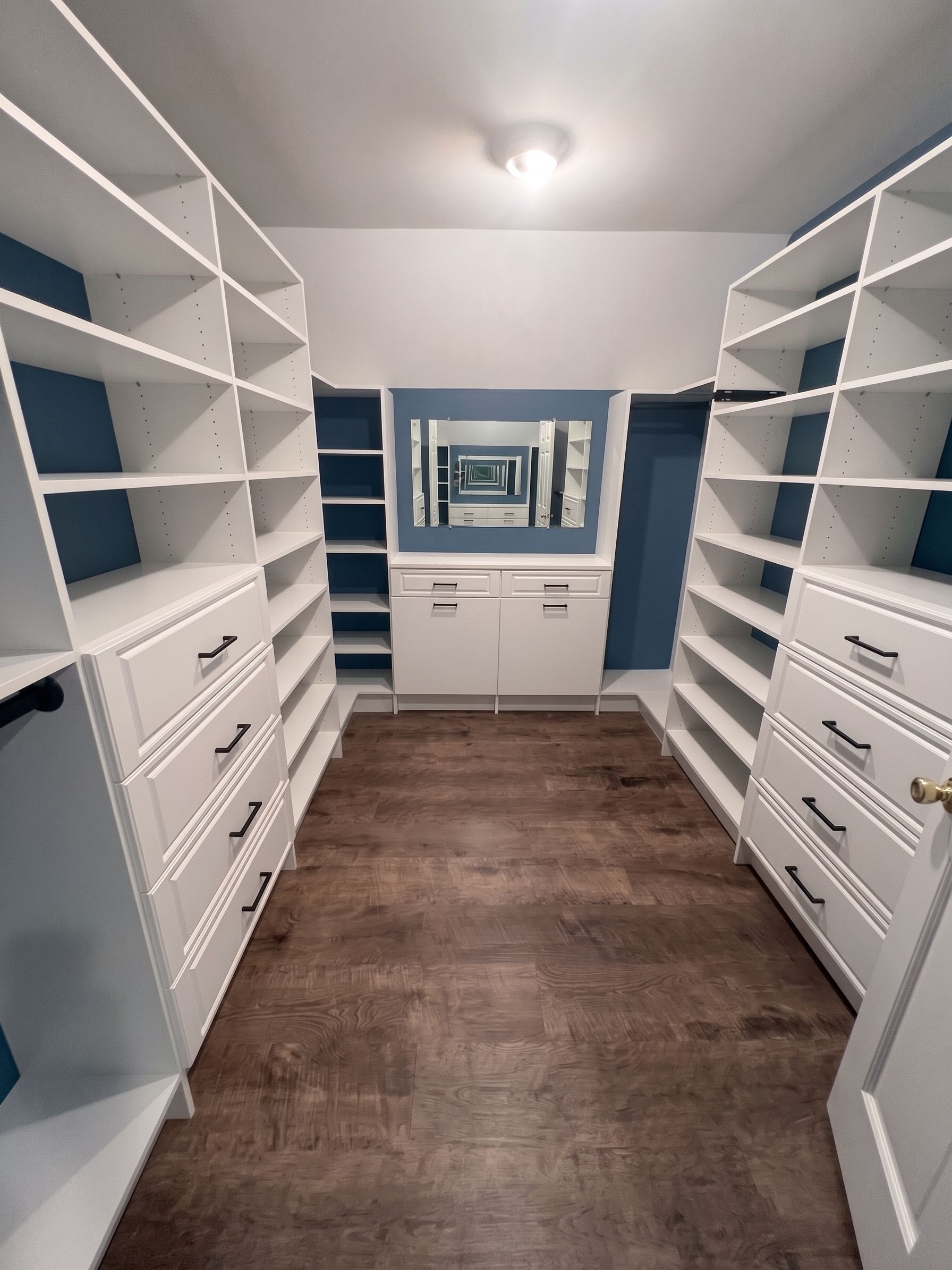 Walk-in closet with white shelves, drawers, and a vanity mirror over dark wood flooring